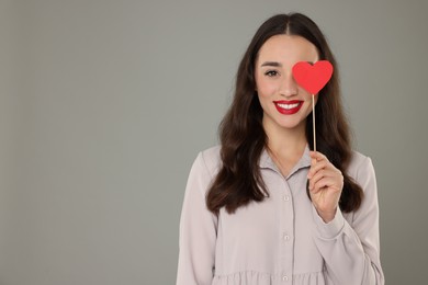 Beautiful young woman covering her eye with paper heart on grey background, space for text