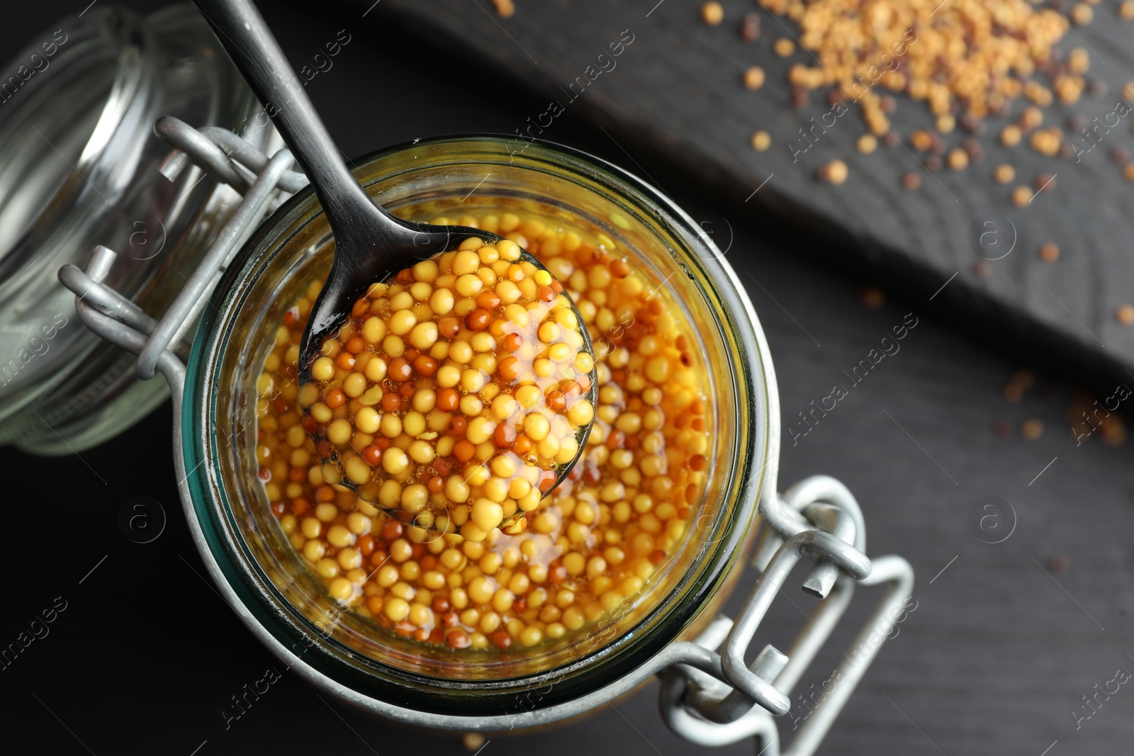 Photo of Whole grain mustard and spoon in jar on black wooden table, top view