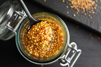 Whole grain mustard and spoon in jar on black wooden table, top view