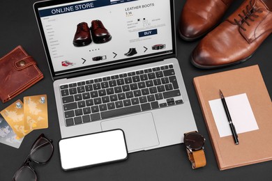 Photo of Online store website on laptop screen. Computer, smartphone, stationery, credit cards, men's shoes and accessories on black background, above view