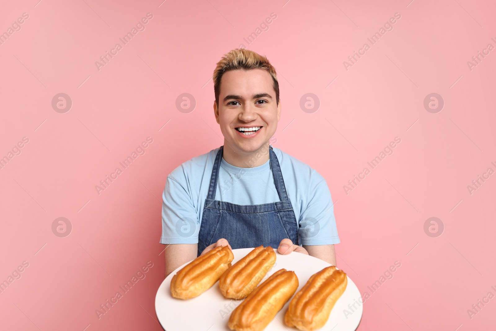 Photo of Portrait of happy confectioner holding plate with delicious eclairs on pink background