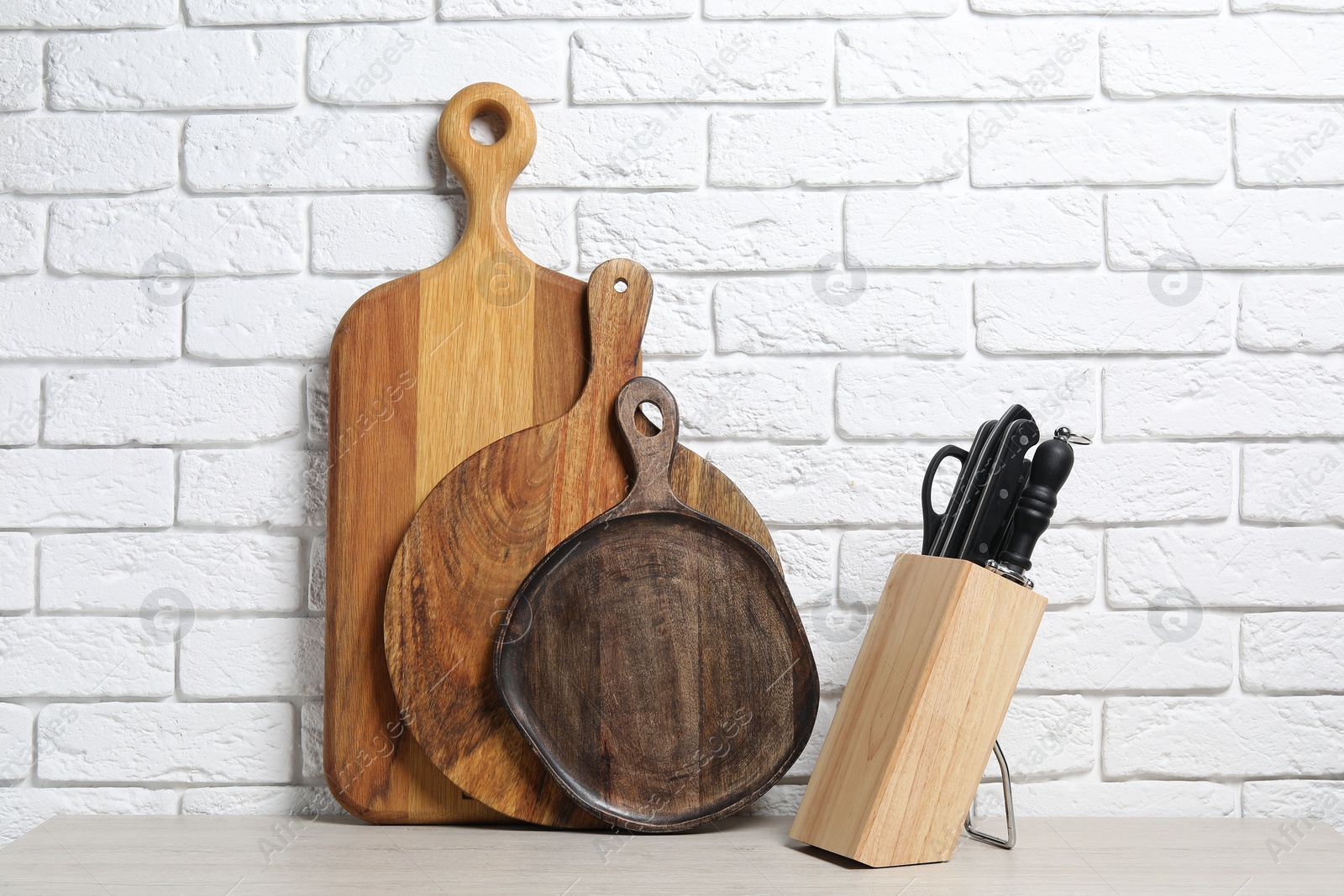 Photo of Different wooden cutting boards and knives in holder on light table near white brick wall