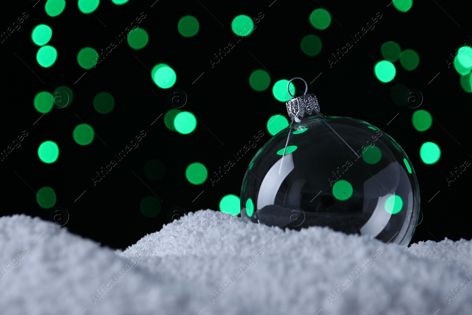 Photo of Transparent Christmas ball on snow against blurred fairy lights, space for text