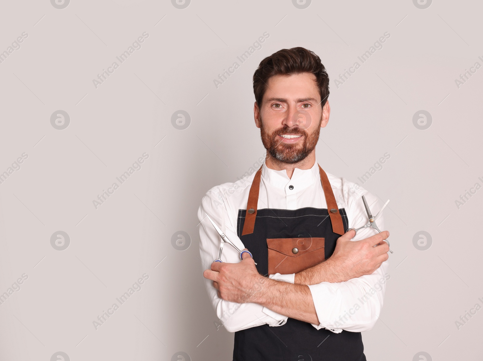 Photo of Smiling hairdresser wearing apron holding scissors on light grey background, space for text