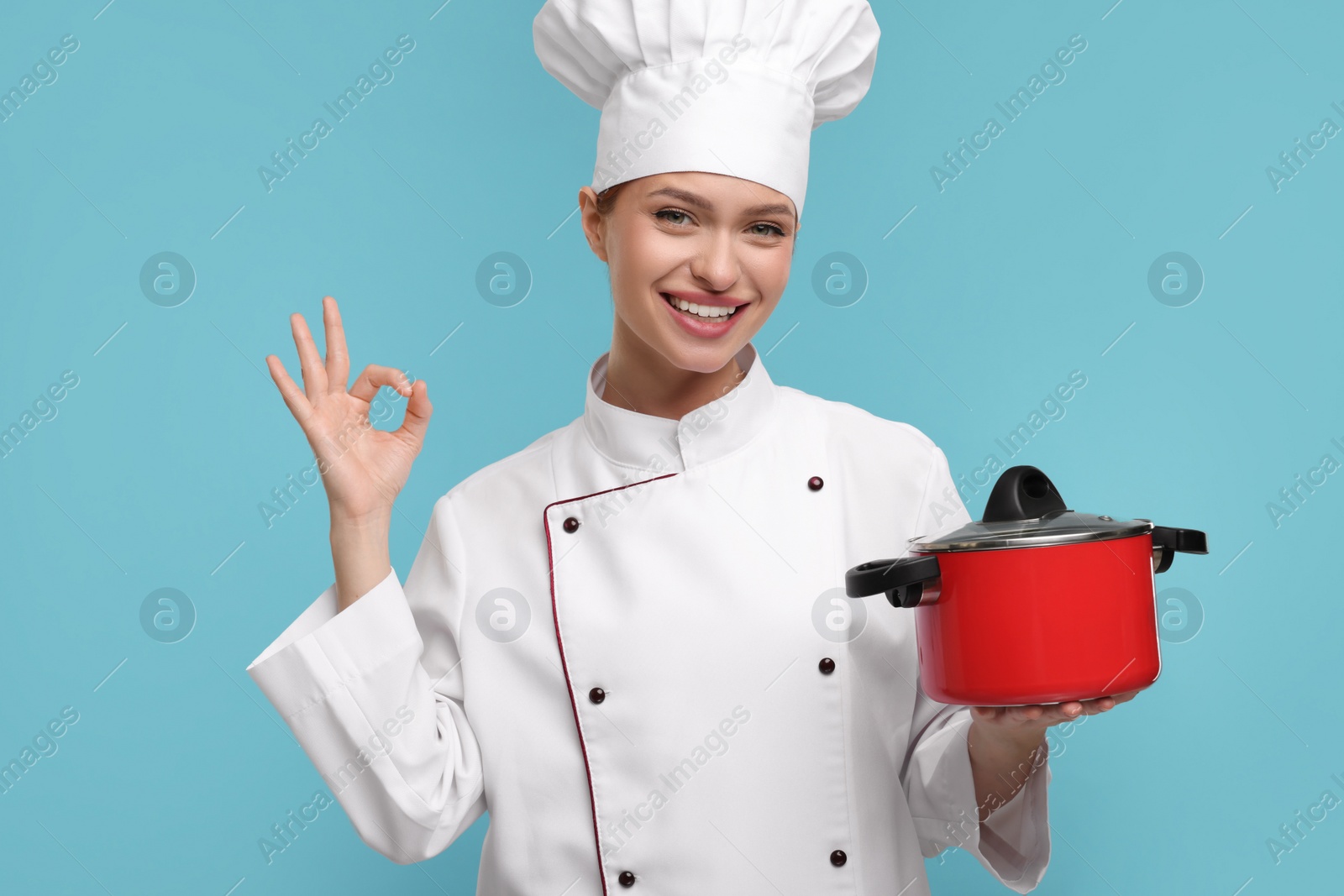 Photo of Happy chef in uniform holding cooking pot and showing OK gesture on light blue background