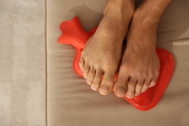 Photo of Man warming feet with hot water bottle on sofa, closeup