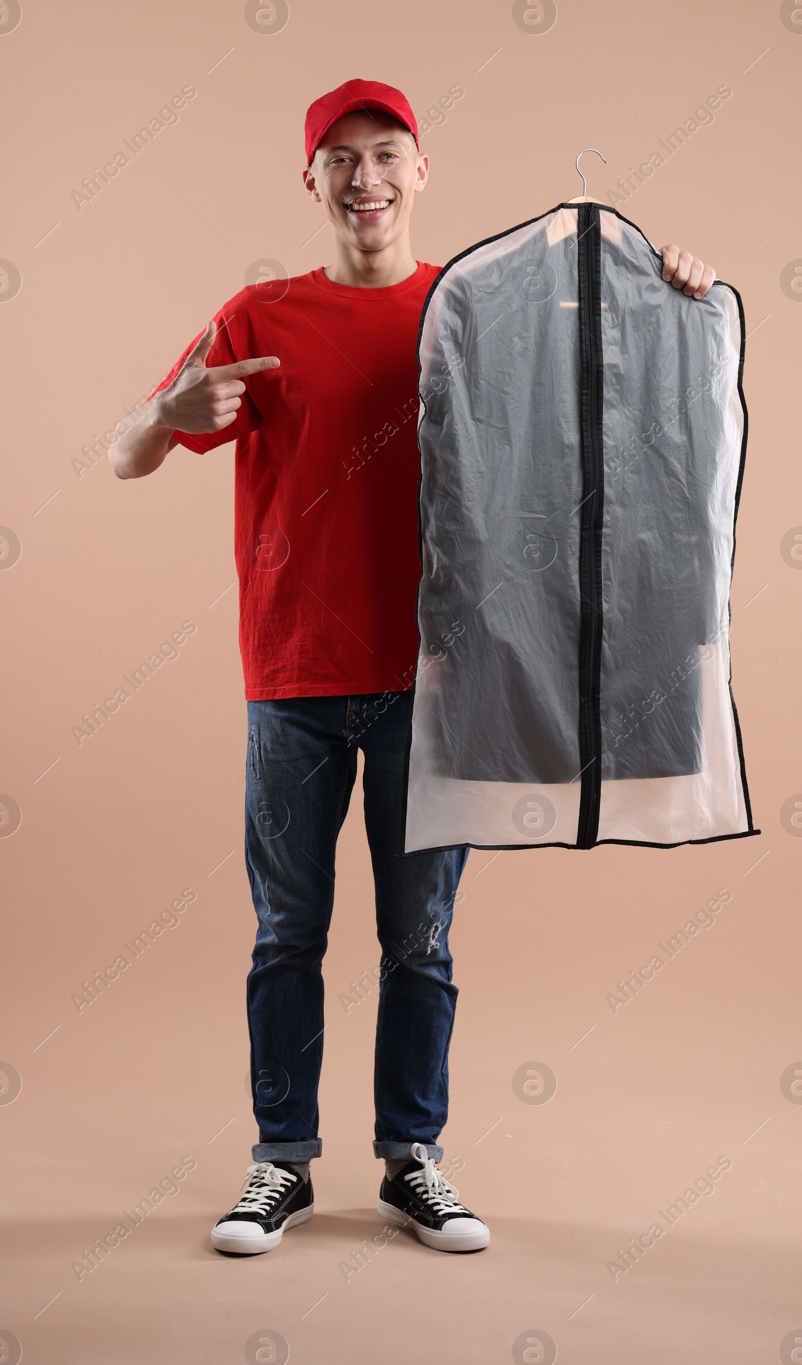 Photo of Dry-cleaning delivery. Happy courier holding garment cover with clothes on beige background