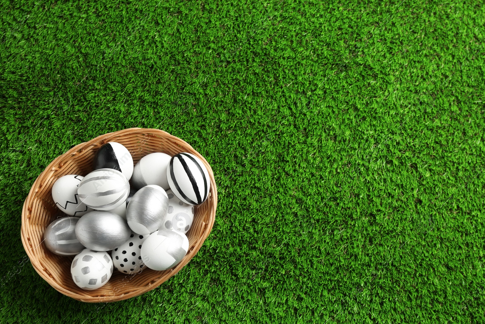 Photo of Wicker basket of painted Easter eggs on green lawn, top view with space for text