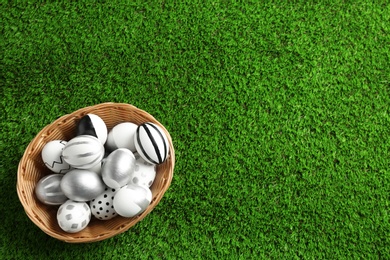 Wicker basket of painted Easter eggs on green lawn, top view with space for text