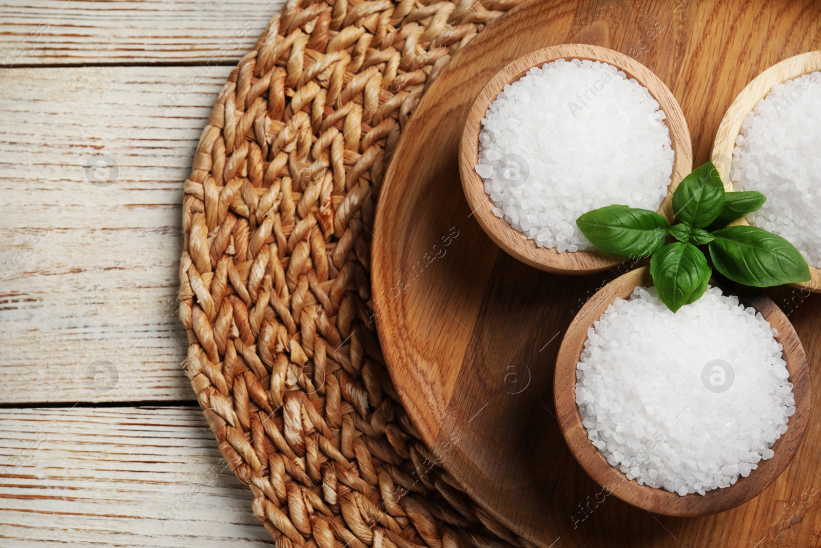 Photo of Bowls of natural sea salt and wicker mat on white wooden table, flat lay. Space for text