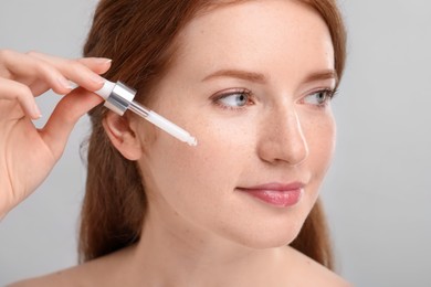 Beautiful woman with freckles applying cosmetic serum onto her face against grey background, closeup