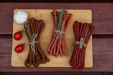 Photo of Bundles of delicious kabanosy with rosemary, tomatoes and garlic on wooden table, top view