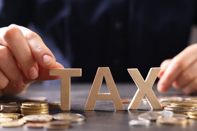 Photo of Woman with word Tax, wooden cubes and coins at grey table, closeup