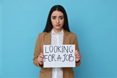 Photo of Young unemployed woman holding sign with phrase Looking For A Job on light blue background