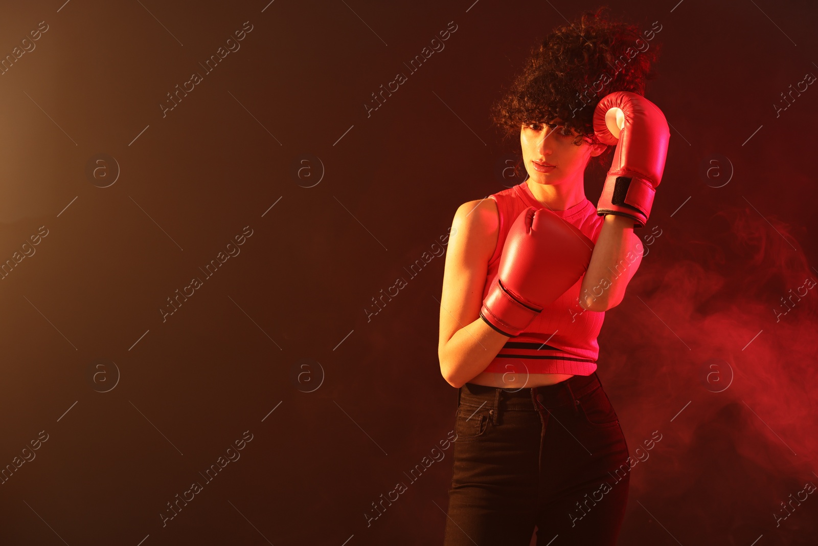 Photo of Beautiful young woman with boxing gloves on color background in neon lights and smoke. Space for text