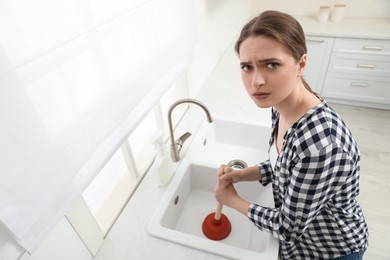 Photo of Upset young woman using plunger to unclog sink drain in kitchen, above view