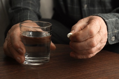 Photo of Senior man holding pill and glass of water at table, closeup