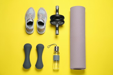 Photo of Exercise mat, dumbbells, bottle of water, ab roller and shoes on yellow background, flat lay