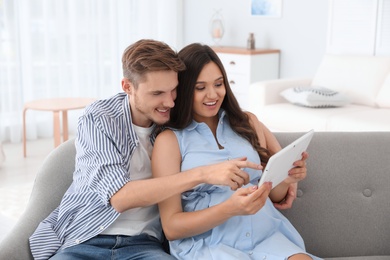 Photo of Young man and his pregnant wife using tablet computer on sofa at home