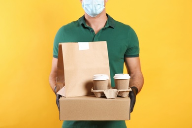 Photo of Courier in medical mask holding packages with takeaway food and drinks on yellow background, closeup. Delivery service during quarantine due to Covid-19 outbreak