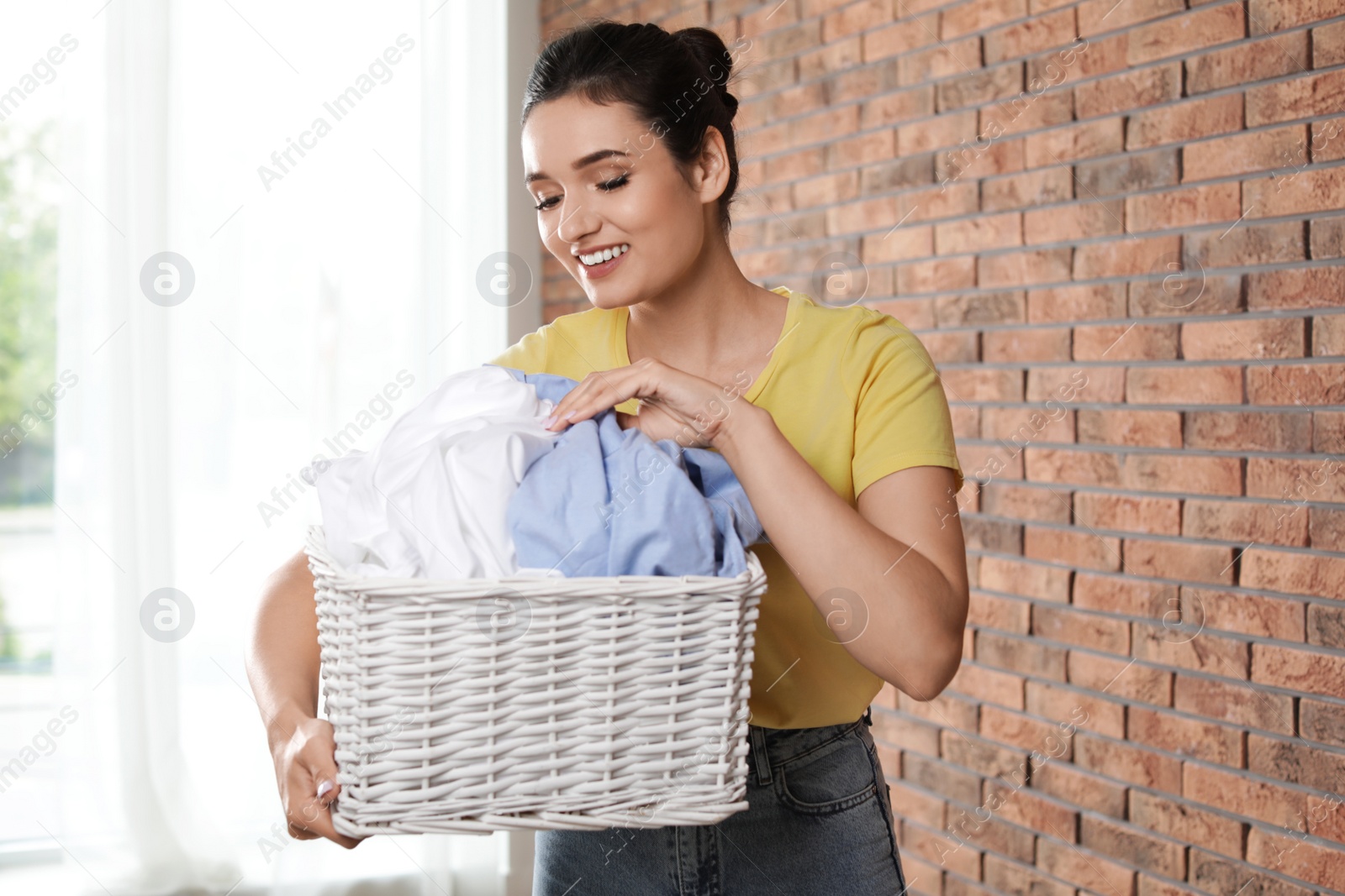 Photo of Young woman with basket full of clean laundry near brick wall indoors