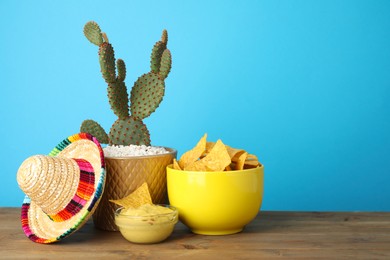 Photo of Mexican sombrero hat, cactus, nachos chips and guacamole in bowls on wooden table against light blue background, space for text