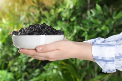 Woman holding bowl of fresh ripe black mulberries on blurred natural background, closeup