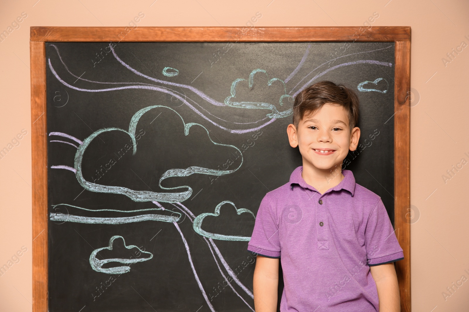 Photo of Cute little child standing at blackboard with chalk drawn sky and clouds