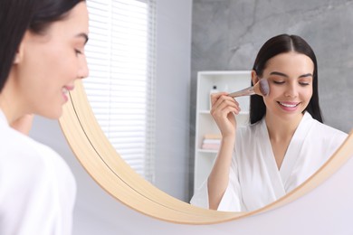 Photo of Beautiful young woman applying makeup with brush near mirror in bathroom