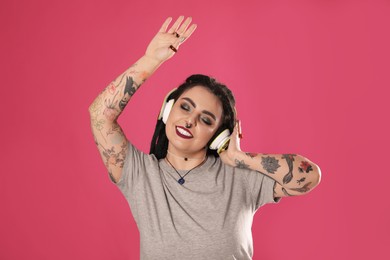 Photo of Beautiful young woman with tattoos on arms, nose piercing and dreadlocks listening to music against pink background