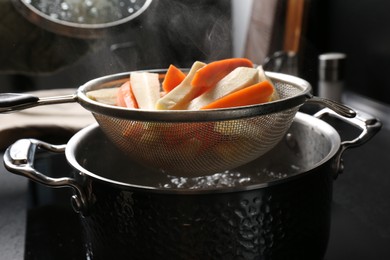 Sieve with cut parsnips and carrots over pot of boiling water in kitchen, closeup