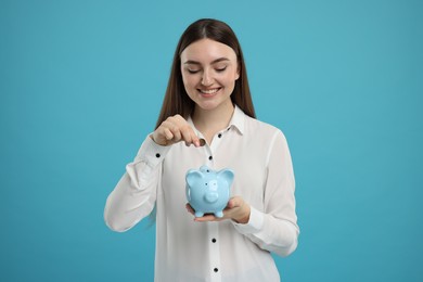 Happy woman putting coin into piggy bank on light blue background