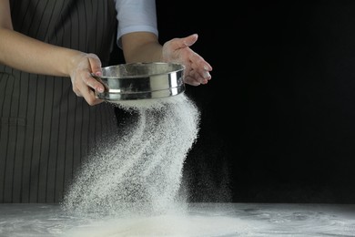 Photo of Woman sieving flour at table against black background, closeup. Space for text