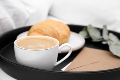 Tray with tasty croissant, cup of coffee and envelope on white bed, closeup