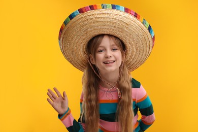 Photo of Cute girl in Mexican sombrero hat waving hello on orange background