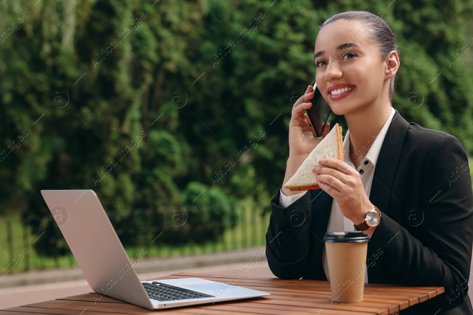 Photo of Happy businesswoman with sandwich talking on smartphone while having lunch at wooden table outdoors