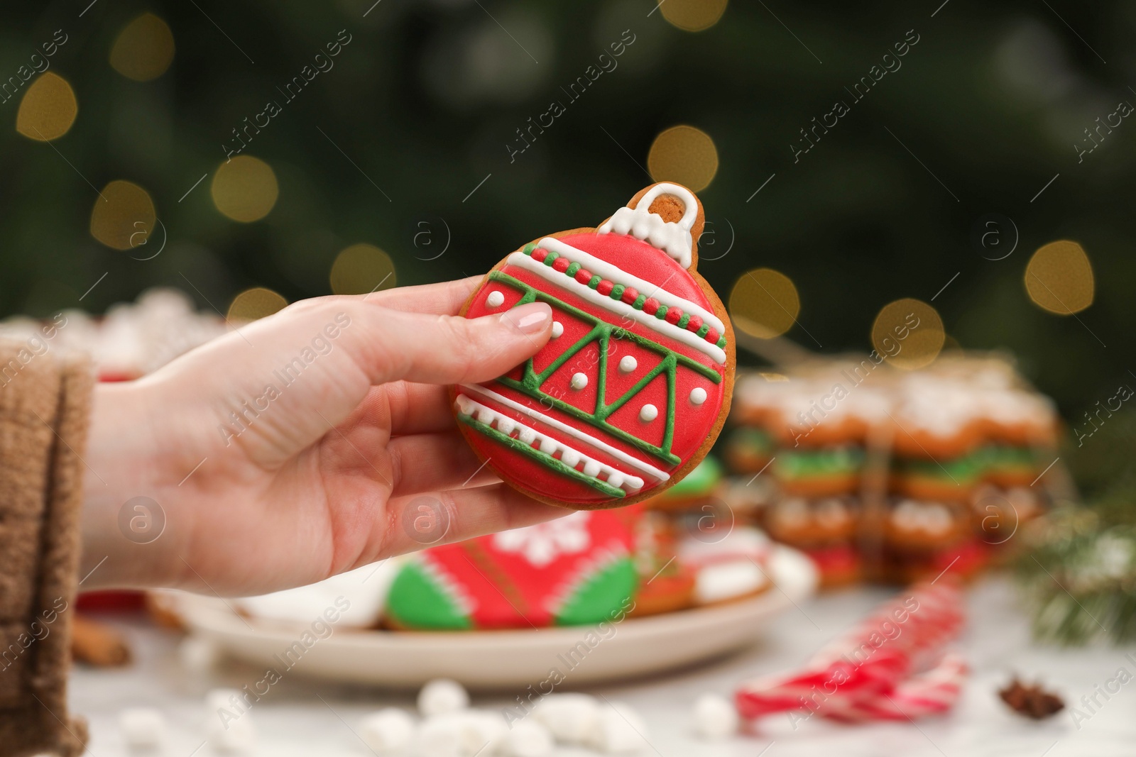 Photo of Woman with decorated cookie at table against blurred Christmas lights, closeup