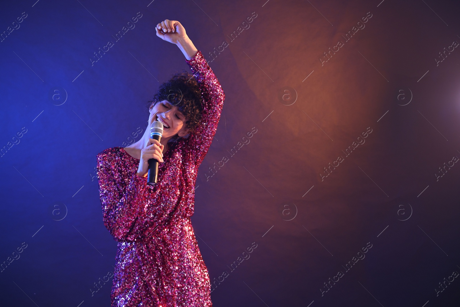 Photo of Beautiful young woman with microphone singing on color background in neon lights. Space for text