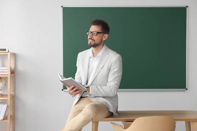 Photo of Happy teacher with book at desk in classroom