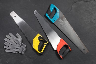 Saws with colorful handles and gloves on black textured background, flat lay