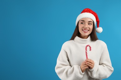 Young woman in beige sweater and Santa hat holding candy cane on light blue background, space for text. Celebrating Christmas