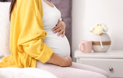 Photo of Young pregnant woman sitting on bed and touching her belly at home, closeup