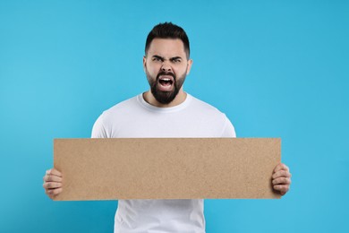 Angry man holding blank cardboard banner on light blue background, space for text