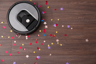 Modern robotic vacuum cleaner removing confetti from wooden floor, top view. Space for text
