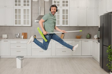 Photo of Enjoying cleaning. Man in headphones jumping with mop in kitchen