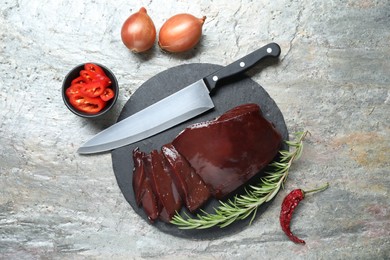 Photo of Cut raw beef liver with rosemary, chili pepper, onions and knife on grey table, flat lay