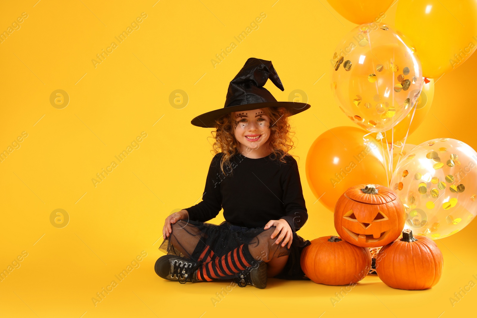 Photo of Cute little girl with pumpkins and balloons wearing Halloween costume on yellow background