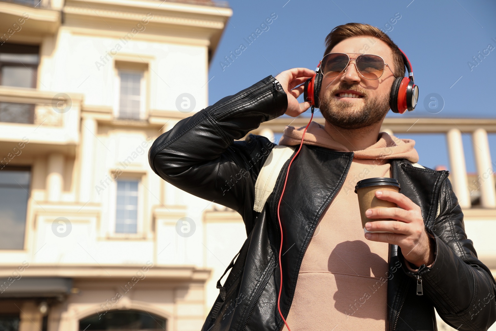 Photo of Happy young man with coffee and headphones listening to music on city street