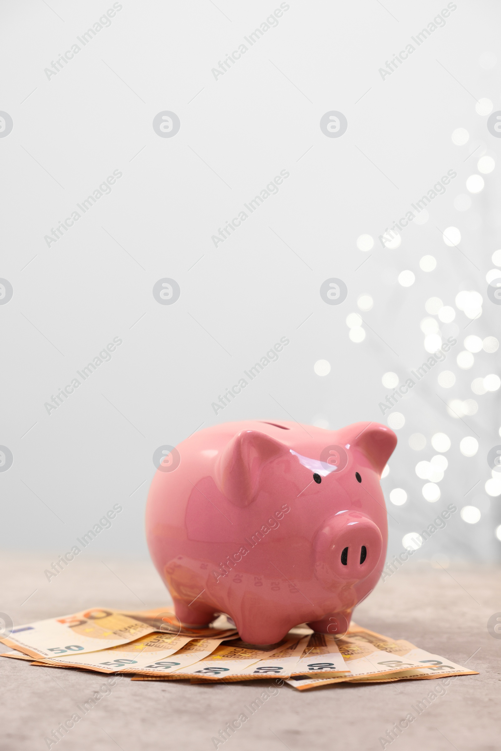 Photo of Piggy bank with euro banknotes on grey table against blurred lights, space for text