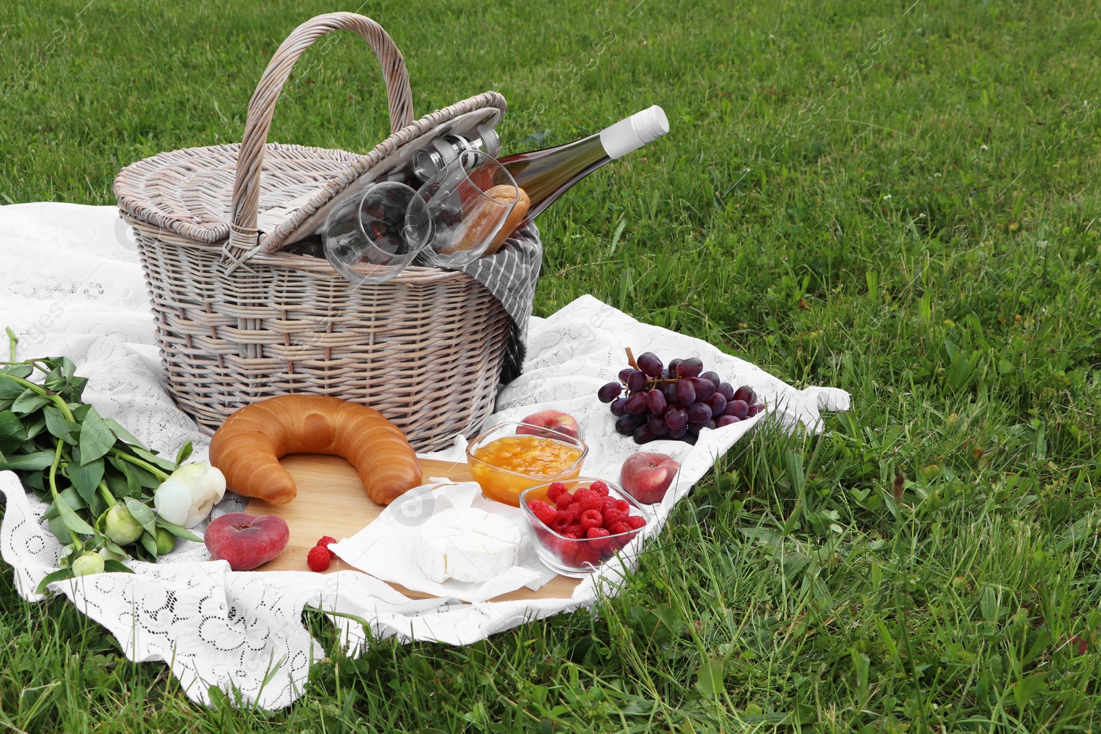 Photo of Picnic blanket with tasty food, flowers, basket and cider on green grass outdoors